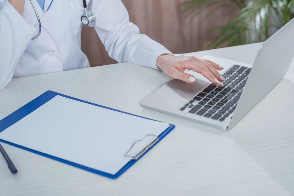 Ärztin mit Stethoskop arbeitet am Laptop, Klemmbrett mit Dokumenten liegt daneben